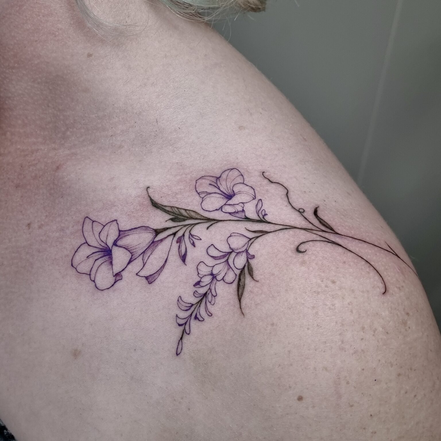 Floral tattoo on shoulder with purple flowers and green leaves.