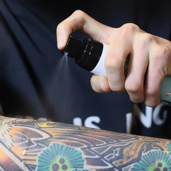 Hand spraying lotion on a colorful tattooed arm.